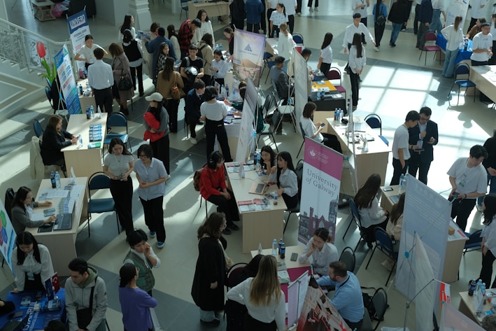 Numerous individuals are gathered in a spacious, well-lit hall, interacting around various booths and tables. Some tables are adorned with informational posters and brochures. People are engaged in discussions, examining flyers, and working on laptops. The setting appears to be a university or career fair, with banners and promotional materials visible in the background.