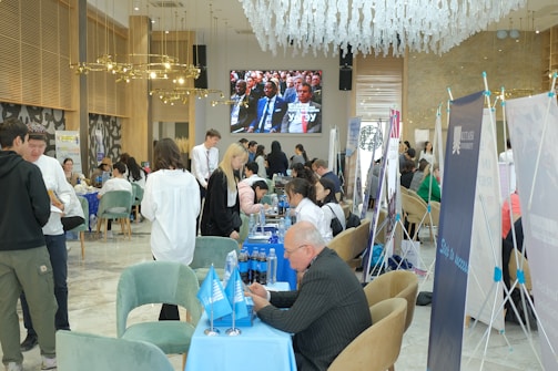A busy room with people seated at tables, engaged in conversation and activities typical of a fair or conference. Banners and flags are displayed prominently, and a large screen shows a man speaking to an audience. The interior is elegant with modern decor, featuring chandeliers and stylish furniture.