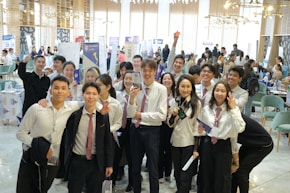 A group of young people in formal attire, predominantly white shirts and ties, gathered indoors with some holding brochures or documents. They are smiling and appear to be posing for a group photo. The background is a busy, well-lit room with tables, chairs, and people engaged in various activities, possibly a conference or fair.