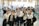 A group of young people in formal attire, predominantly white shirts and ties, gathered indoors with some holding brochures or documents. They are smiling and appear to be posing for a group photo. The background is a busy, well-lit room with tables, chairs, and people engaged in various activities, possibly a conference or fair.