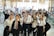 A group of young people in formal attire, predominantly white shirts and ties, gathered indoors with some holding brochures or documents. They are smiling and appear to be posing for a group photo. The background is a busy, well-lit room with tables, chairs, and people engaged in various activities, possibly a conference or fair.