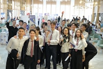 A group of young people in formal attire, predominantly white shirts and ties, gathered indoors with some holding brochures or documents. They are smiling and appear to be posing for a group photo. The background is a busy, well-lit room with tables, chairs, and people engaged in various activities, possibly a conference or fair.