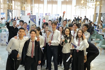 A group of young people in formal attire, predominantly white shirts and ties, gathered indoors with some holding brochures or documents. They are smiling and appear to be posing for a group photo. The background is a busy, well-lit room with tables, chairs, and people engaged in various activities, possibly a conference or fair.