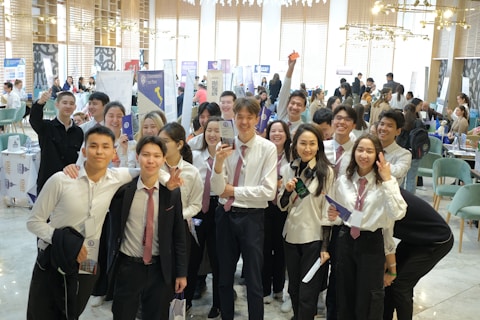 A group of young people in formal attire, predominantly white shirts and ties, gathered indoors with some holding brochures or documents. They are smiling and appear to be posing for a group photo. The background is a busy, well-lit room with tables, chairs, and people engaged in various activities, possibly a conference or fair.