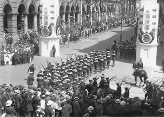 A large procession featuring rows of uniformed soldiers marching in formation along a street lined with onlookers. The scene includes decorated columns and a building with arched windows. The crowd surrounds the parade, and there are several people on horseback.