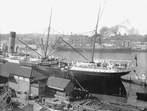 A bustling port scene with several large steamships docked along the waterfront. The ships have tall masts and smokestacks, and there are various buildings and warehouses in the background. Smoke rises from some of the ships, indicating activity. On the dock, people and horse-drawn carts are visible, suggesting industrial commerce. The setting appears to be in the early 20th century.