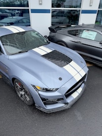 A sleek, high-performance sports car with a blue body and white racing stripes is parked next to a similar vehicle. The car features aerodynamic design elements, a prominent hood vent, and intricate wheel designs. The setting appears to be outside an automotive showroom or dealership, with reflections visible in the glass windows.