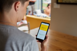 A person is holding a smartphone displaying a graph with a numerical value on a yellow background. The scene takes place indoors at a table, with other people blurred in the background. The focus is on the phone screen, which shows a line graph and the number 11.7.