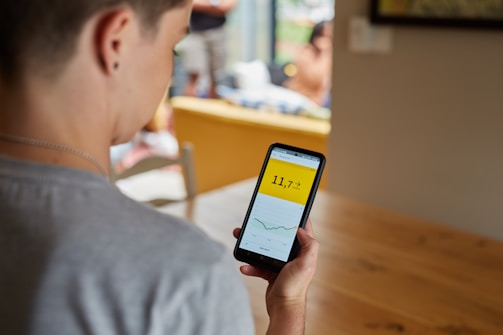 A person is holding a smartphone displaying a graph with a numerical value on a yellow background. The scene takes place indoors at a table, with other people blurred in the background. The focus is on the phone screen, which shows a line graph and the number 11.7.