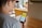 A person is holding a smartphone displaying a graph with a numerical value on a yellow background. The scene takes place indoors at a table, with other people blurred in the background. The focus is on the phone screen, which shows a line graph and the number 11.7.