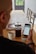 A person is sitting at a wooden dining table holding a smartphone displaying an application screen. On the table, there is a French press with coffee, a mug filled with coffee, a bowl containing cookies, and a magazine.