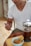 A person is seated and administering an injection into their thigh. They are wearing a white t-shirt and khaki shorts. In the foreground, there is a French press filled with coffee and a small bowl containing breadsticks on a wooden table.