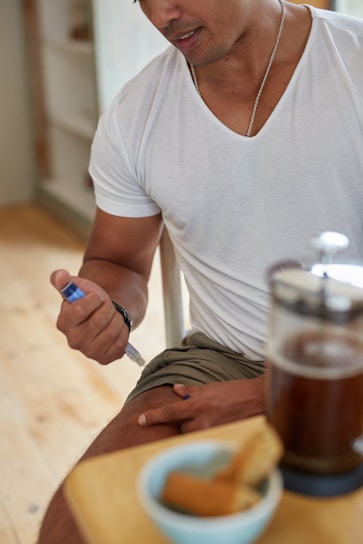 A person is seated and administering an injection into their thigh. They are wearing a white t-shirt and khaki shorts. In the foreground, there is a French press filled with coffee and a small bowl containing breadsticks on a wooden table.