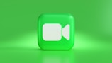 A 3D, square-shaped icon with rounded edges features a camera symbol in white against a vibrant green background. The icon creates a visually striking effect with its glossy finish and the soft lighting surrounding it.