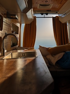The interior of a van features wooden paneling and a sleek countertop with a metallic sink. Yellow curtains frame the rear opening, revealing a serene landscape in the distance. Cozy cushions and bedding suggest a comfortable living space.