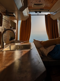The interior of a van features wooden paneling and a sleek countertop with a metallic sink. Yellow curtains frame the rear opening, revealing a serene landscape in the distance. Cozy cushions and bedding suggest a comfortable living space.