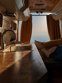 The interior of a van features wooden paneling and a sleek countertop with a metallic sink. Yellow curtains frame the rear opening, revealing a serene landscape in the distance. Cozy cushions and bedding suggest a comfortable living space.
