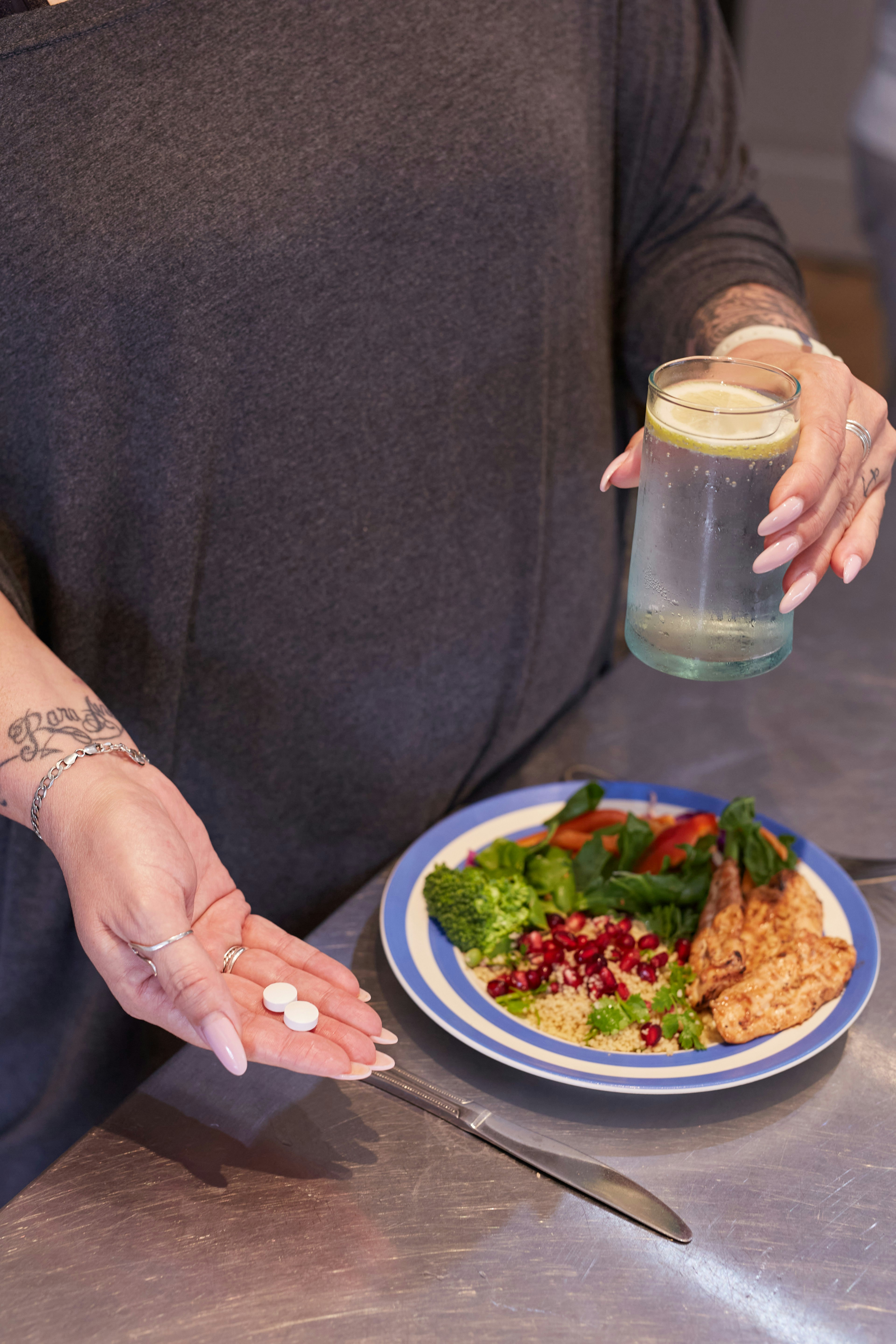 A person holds two white pills in one hand and a glass of water with a lemon slice in the other. On the table in front, there is a plate with a meal consisting of grilled chicken, broccoli, spinach, quinoa, and pomegranate seeds. The person is wearing a dark top and has tattoos on their forearm.