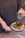 A person holds two white pills in one hand and a glass of water with a lemon slice in the other. On the table in front, there is a plate with a meal consisting of grilled chicken, broccoli, spinach, quinoa, and pomegranate seeds. The person is wearing a dark top and has tattoos on their forearm.
