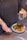 A person holds two white pills in one hand and a glass of water with a lemon slice in the other. On the table in front, there is a plate with a meal consisting of grilled chicken, broccoli, spinach, quinoa, and pomegranate seeds. The person is wearing a dark top and has tattoos on their forearm.