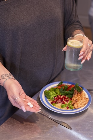 A person holds two white pills in one hand and a glass of water with a lemon slice in the other. On the table in front, there is a plate with a meal consisting of grilled chicken, broccoli, spinach, quinoa, and pomegranate seeds. The person is wearing a dark top and has tattoos on their forearm.