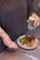 A person holds two white pills in one hand and a glass of water with a lemon slice in the other. On the table in front, there is a plate with a meal consisting of grilled chicken, broccoli, spinach, quinoa, and pomegranate seeds. The person is wearing a dark top and has tattoos on their forearm.