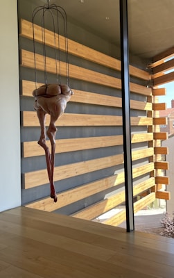 A modern interior with wooden paneling on the walls and a large glass window, showcasing an abstract sculpture hanging from the ceiling. The sculpture resembles a human torso with elongated limbs, crafted in a surreal style. Natural light enters through the window, illuminating the room.