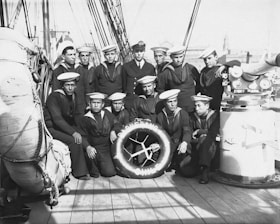 A group of sailors wearing uniforms and caps are gathered on the deck of a ship. The men are arranged in a casual pose around a large life ring. The ship’s deck is equipped with nautical gear such as ropes and machinery, visible in the background and to the sides.