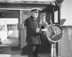 A ship's captain in uniform operates a telegraph on the ship's bridge. The telegraph is a classic brass model with various speed settings visible. Sunlight casts shadows on the deck, enhancing the vintage and maritime atmosphere.