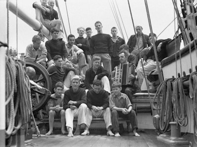 A group of young men and boys, including a man playing an accordion, are seated and standing on the deck of a ship. The ship's rigging, ropes, and wheel are visible. The group appears relaxed, with some individuals looking at the camera and others gazing elsewhere.