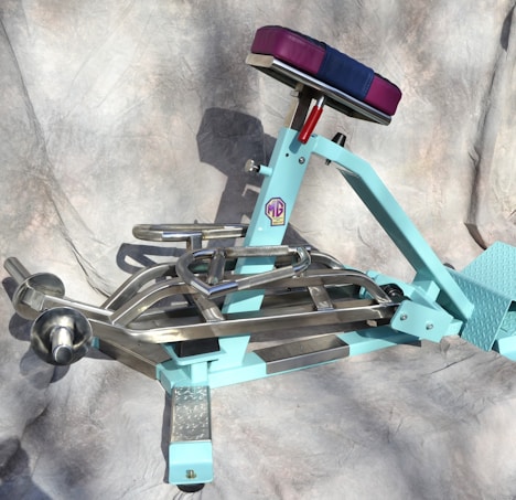 A turquoise exercise machine with metallic handles and a padded seat in contrasting colors. The design appears sturdy with a combination of steel and cushioned parts, designed for workouts. The background is a neutral fabric backdrop, providing a simple yet effective contrast to the machine.