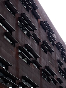 A tall building with a modern architectural design featuring a grid pattern of rectangular windows. The exterior appears to be made of a dark, weathered material, and the structure gives a sense of depth through its repeating geometric elements.