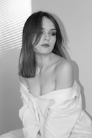 A woman with shoulder-length hair looks to the side, wearing an off-the-shoulder white shirt. The setting is softly lit, with shadows and light creating a pattern from blinds on the wall.