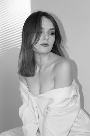 A woman with shoulder-length hair looks to the side, wearing an off-the-shoulder white shirt. The setting is softly lit, with shadows and light creating a pattern from blinds on the wall.