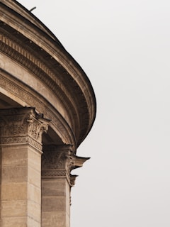 Detailed architectural elements of a classical building with ornate columns and intricate carvings. The image focuses on the elegant curves and detailed stonework of the structure against a plain sky.