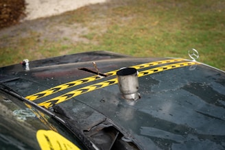 A view of an old, worn-out car hood with two yellow and black striped caution tapes placed diagonally. The hood appears dusty and rusted, with a metal exhaust pipe protruding from it and a Mercedes-Benz emblem visible on the front. The background shows a grassy area with a gravel path.