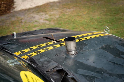A view of an old, worn-out car hood with two yellow and black striped caution tapes placed diagonally. The hood appears dusty and rusted, with a metal exhaust pipe protruding from it and a Mercedes-Benz emblem visible on the front. The background shows a grassy area with a gravel path.