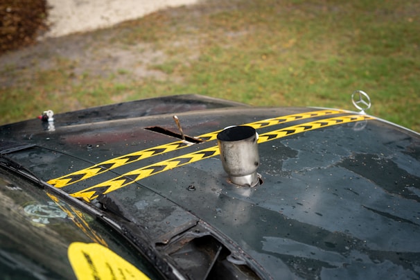 A view of an old, worn-out car hood with two yellow and black striped caution tapes placed diagonally. The hood appears dusty and rusted, with a metal exhaust pipe protruding from it and a Mercedes-Benz emblem visible on the front. The background shows a grassy area with a gravel path.
