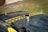 A view of an old, worn-out car hood with two yellow and black striped caution tapes placed diagonally. The hood appears dusty and rusted, with a metal exhaust pipe protruding from it and a Mercedes-Benz emblem visible on the front. The background shows a grassy area with a gravel path.