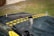 A view of an old, worn-out car hood with two yellow and black striped caution tapes placed diagonally. The hood appears dusty and rusted, with a metal exhaust pipe protruding from it and a Mercedes-Benz emblem visible on the front. The background shows a grassy area with a gravel path.