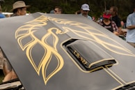 A car hood featuring a large, stylized golden phoenix decal. The foreground includes part of the car's scoop labeled 'TA-427'. Several people are gathered around, some wearing hats and caps, suggesting an outdoor event.