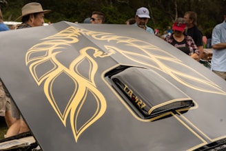 A car hood featuring a large, stylized golden phoenix decal. The foreground includes part of the car's scoop labeled 'TA-427'. Several people are gathered around, some wearing hats and caps, suggesting an outdoor event.