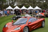 A red sports car is prominently displayed on a green lawn, surrounded by people walking and admiring the vehicles. Behind the car are several white tents with groups of people gathered, forming a backdrop. The scene is lively, suggesting an outdoor car show or exhibition.