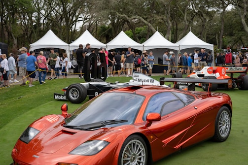 A red sports car is prominently displayed on a green lawn, surrounded by people walking and admiring the vehicles. Behind the car are several white tents with groups of people gathered, forming a backdrop. The scene is lively, suggesting an outdoor car show or exhibition.