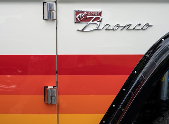 A close-up view of a vehicle door featuring horizontal stripes in shades of red and orange with chrome hinges and a shiny black fender. The emblem has the words 'Sport Bronco' with a stylized horse, positioned above the stripes.