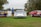 Three vintage race cars are parked on a grassy field, with people walking around and viewing them. The cars display vibrant racing decals and numbers, and a sign with information about the vehicles is visible in front of one car. Trees surround the area, and the sky is overcast.
