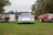 Three vintage race cars are parked on a grassy field, with people walking around and viewing them. The cars display vibrant racing decals and numbers, and a sign with information about the vehicles is visible in front of one car. Trees surround the area, and the sky is overcast.