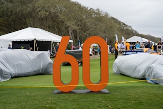 An outdoor event displays vehicles covered with protective covers on a grassy area. A large orange '60' stands prominently in the foreground. People are mingling in the background, with several tents alongside a wooded area. The setting appears casual with a mix of adults and some children.