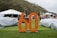 An outdoor event displays vehicles covered with protective covers on a grassy area. A large orange '60' stands prominently in the foreground. People are mingling in the background, with several tents alongside a wooded area. The setting appears casual with a mix of adults and some children.
