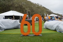 An outdoor event displays vehicles covered with protective covers on a grassy area. A large orange '60' stands prominently in the foreground. People are mingling in the background, with several tents alongside a wooded area. The setting appears casual with a mix of adults and some children.